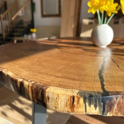 Studio Dekoracji Wnętrz Forrest - Okrągły blat drewniany z żywicą, naturalna krawędź, na metalowej nodze. W tle schody i żółte kwiaty w wazonie. Słoneczne światło podkreśla unikalny charakter drewna.