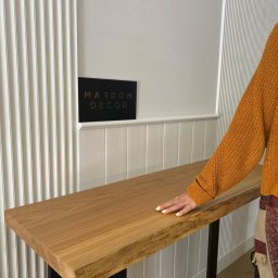 Studio Dekoracji Wnętrz Forrest - Długi, wąski blat drewniany z naturalną krawędzią, wsparty na czarnych, metalowych nogach. Kobieta w pomarańczowym swetrze dotyka blatu. W tle dekoracyjna ściana.