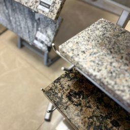 Studio Dekoracji Wnętrz Forrest - Ekspozycja próbek blatów z konglomeratu na metalowym stojaku. Widoczne różne wzory i kolory kamienia, od jasnych szarości po ciemne brązy z czarnymi akcentami.