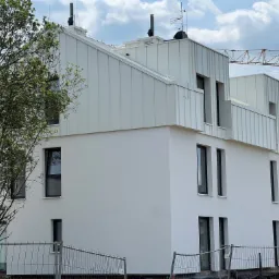 Nowoczesny, biały budynek z dachem z blachy trapezowej, widok z dołu. Na dachu widoczne kominy i anteny. W tle fragment żurawia budowlanego i błękitne niebo.