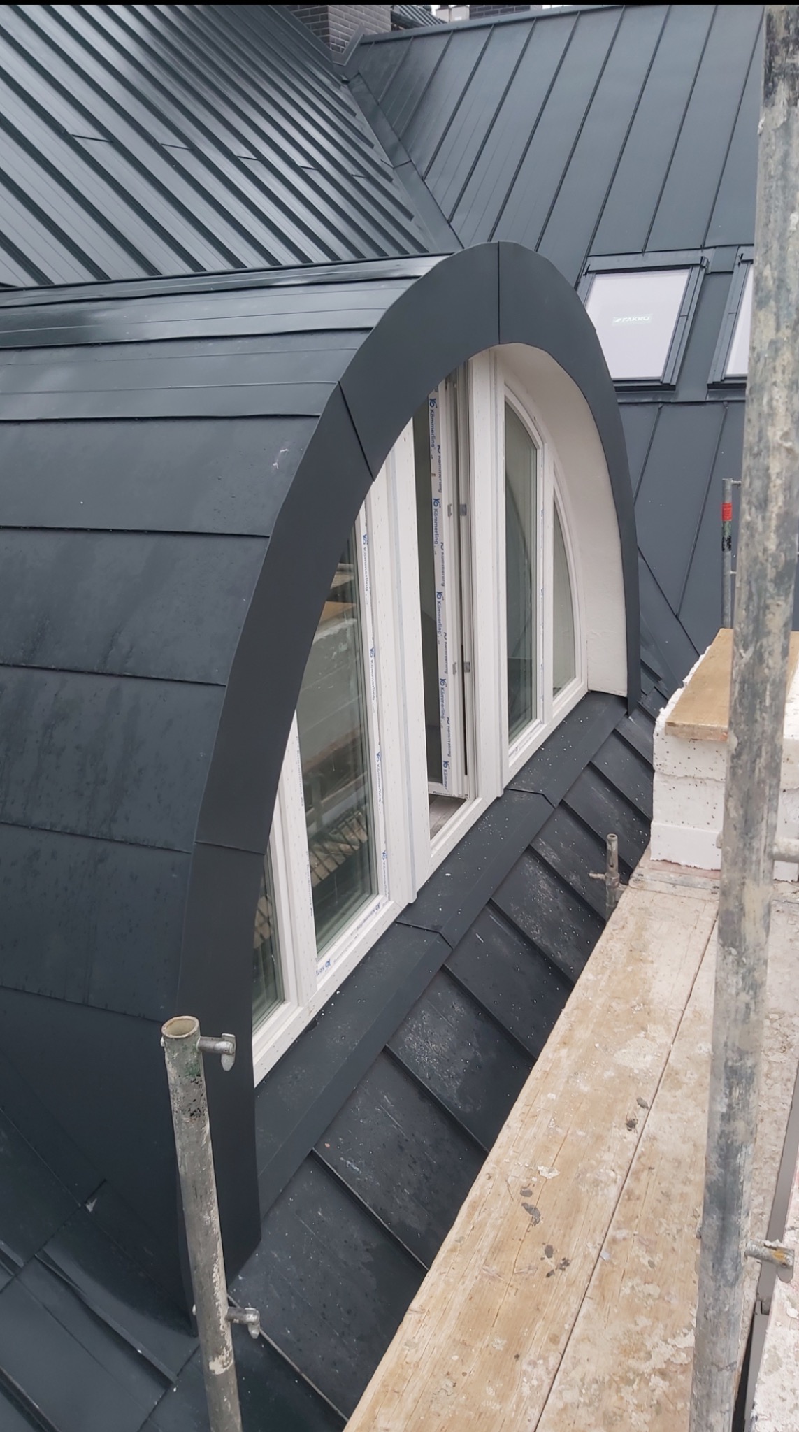 Grafitowy dach z blachy na rąbek stojący z oknem w kształcie półkola, widoczne rusztowanie i okna dachowe Fakro