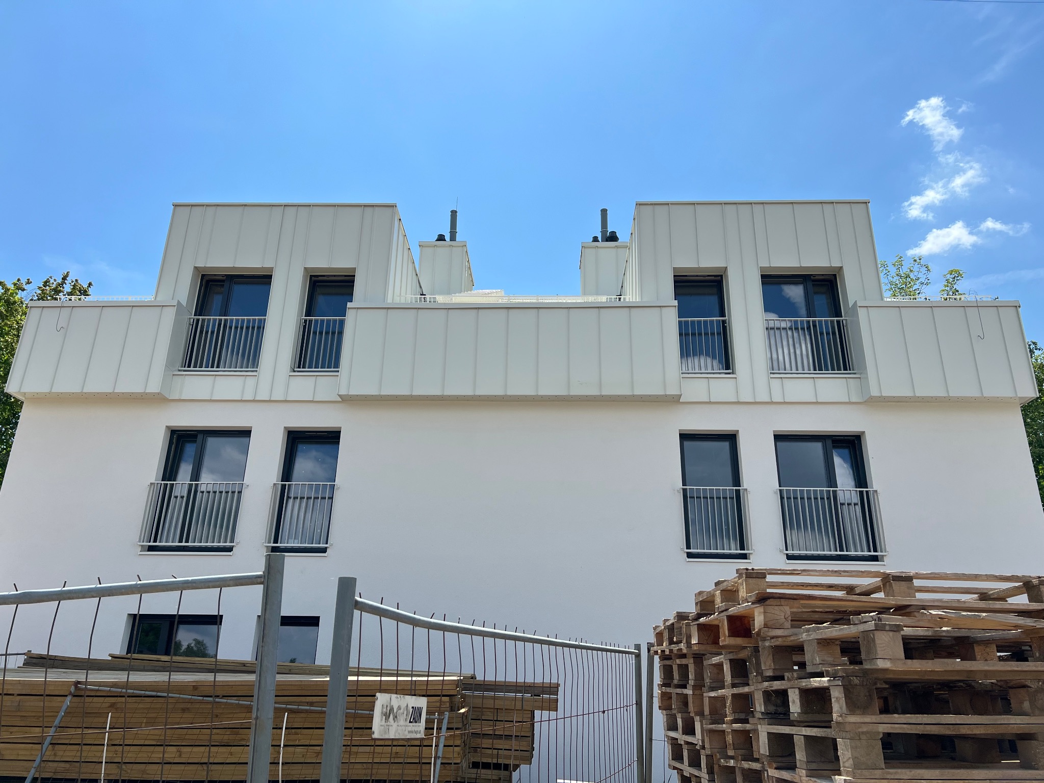 Nowoczesny budynek z białym dachem z blachy trapezowej, balkony z metalowymi barierkami, na pierwszym planie palety i ogrodzenie budowlane.