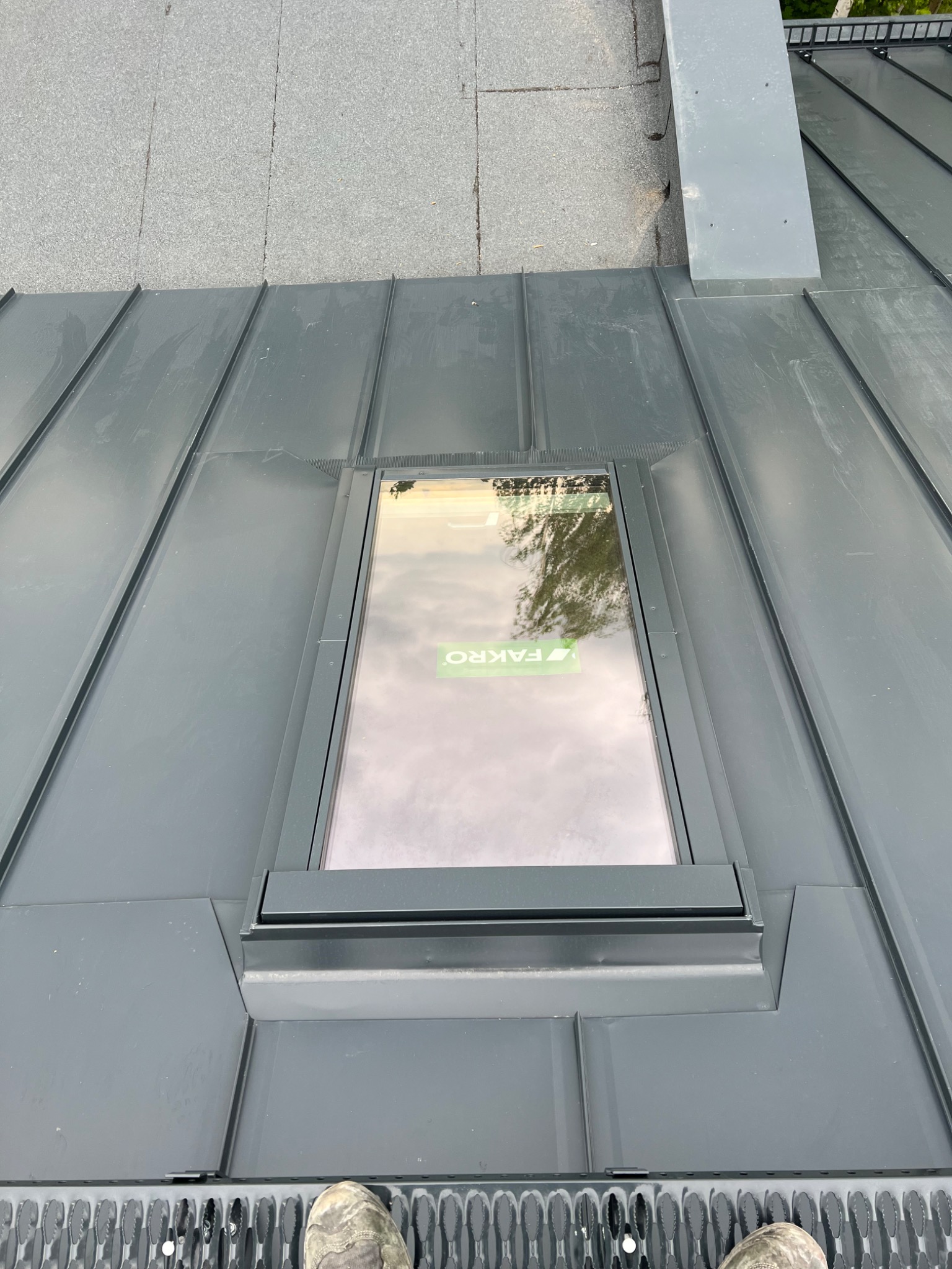 Szary dach z blachy trapezowej z zamontowanym oknem dachowym marki Fakro. Widoczne buty osoby stojącej na dachu, sugerujące prace dekarskie.