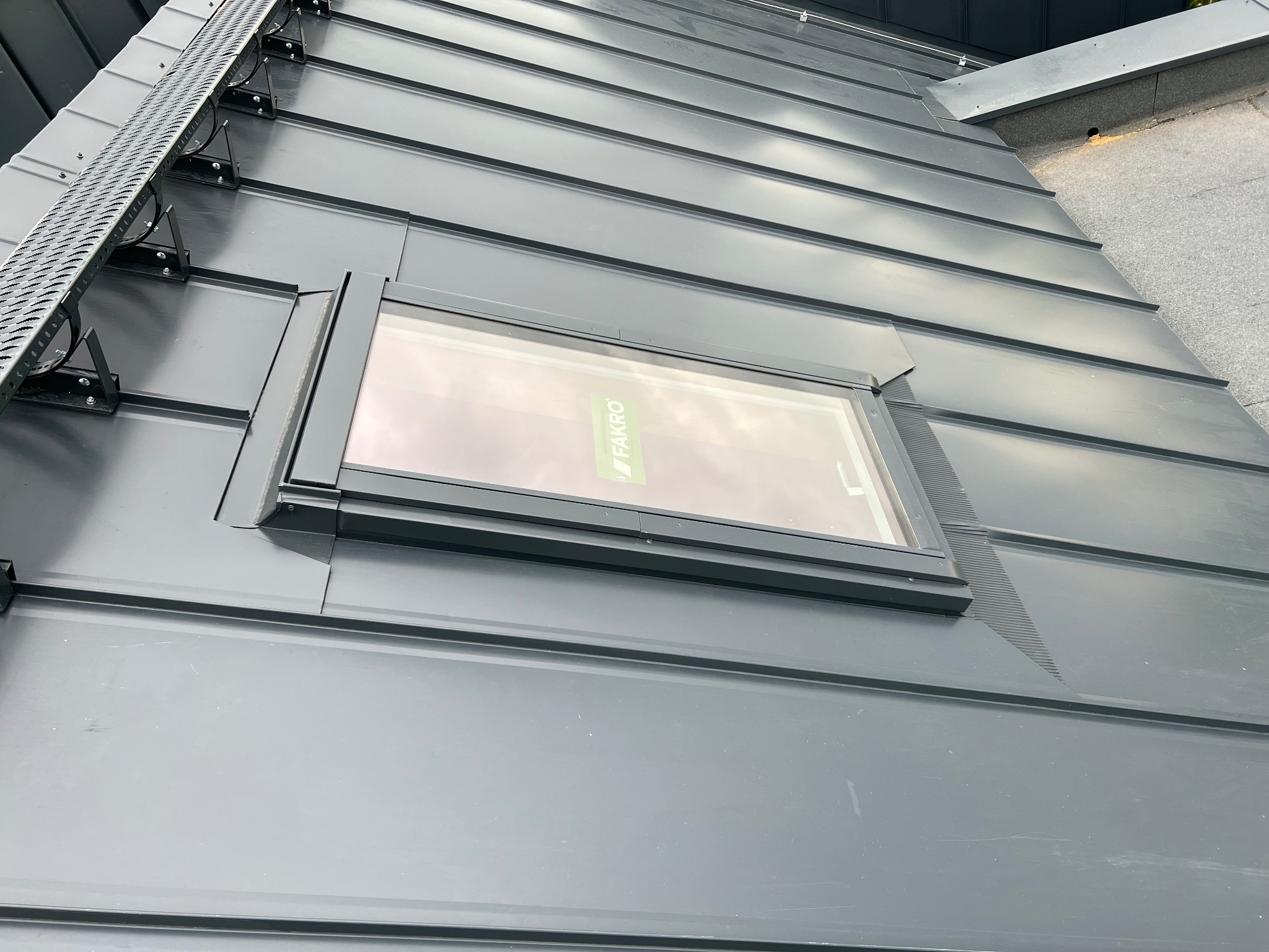 Szary dach z blachy na rąbek stojący z oknem dachowym Fakro i metalową ławą kominiarską. Widok z dołu pod kątem.