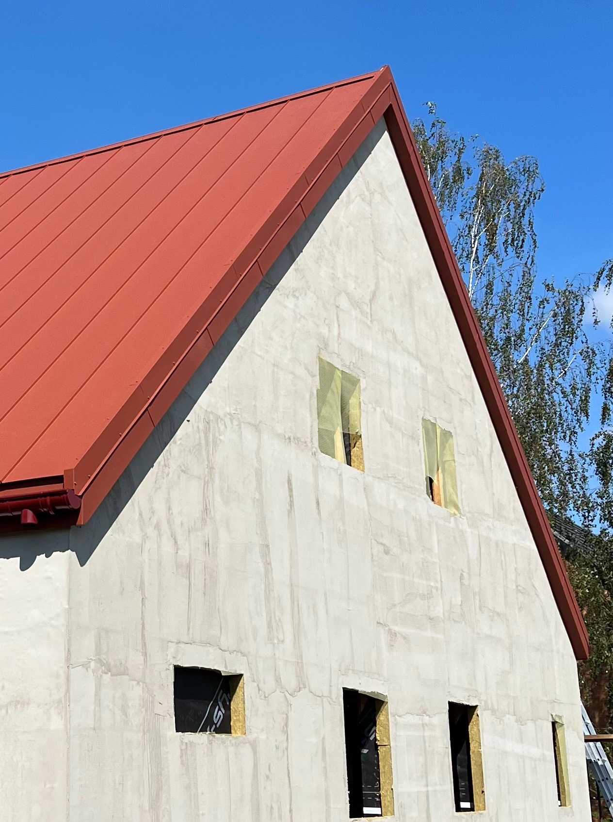 Czerwony, blaszany dach na surowym budynku z oknami zabezpieczonymi folią, widok z dołu na tle błękitnego nieba i drzew.