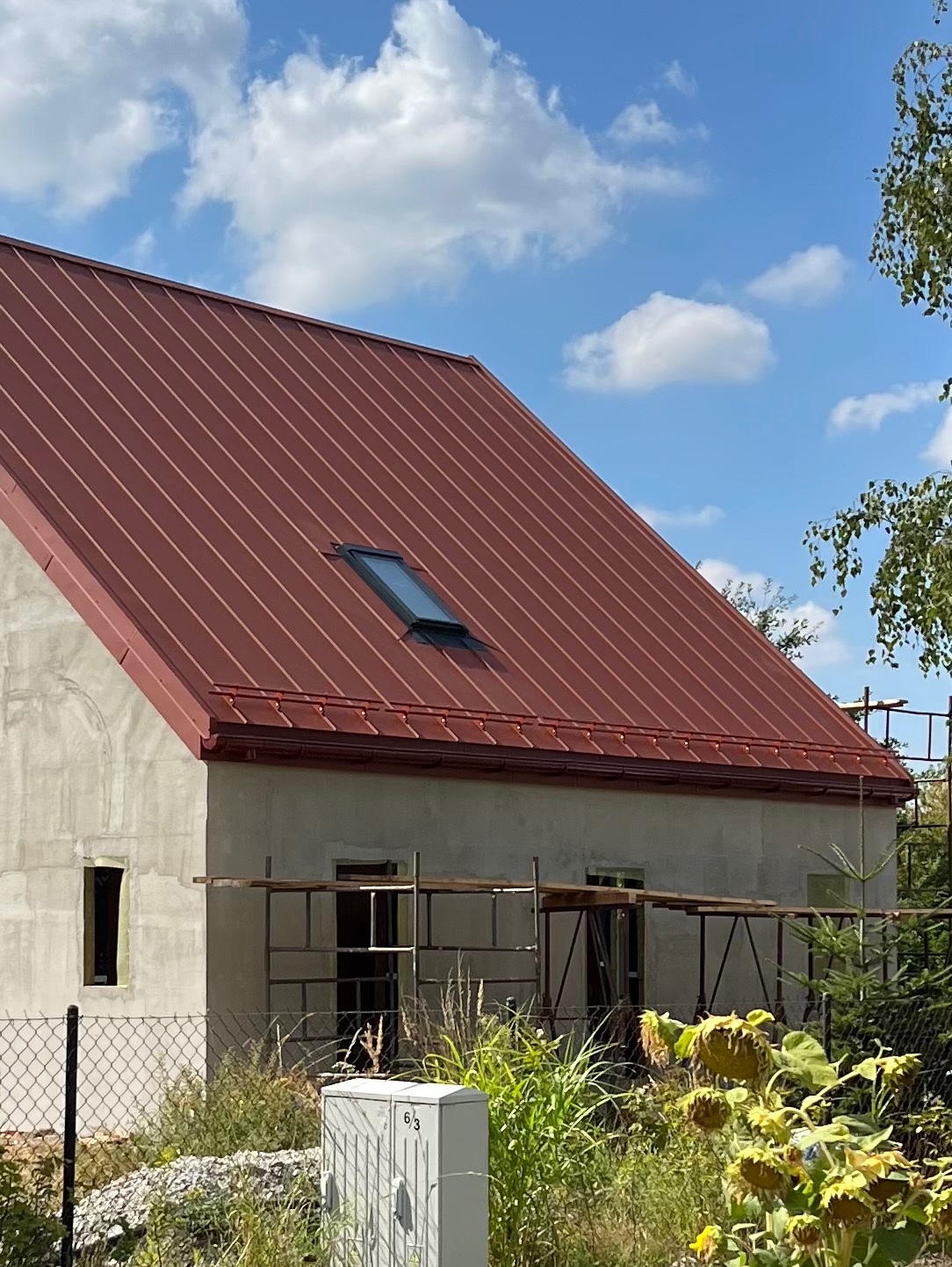 Czerwony dach z blachy na surowym budynku z oknem dachowym i rynnami. Widoczne rusztowania i roślinność.