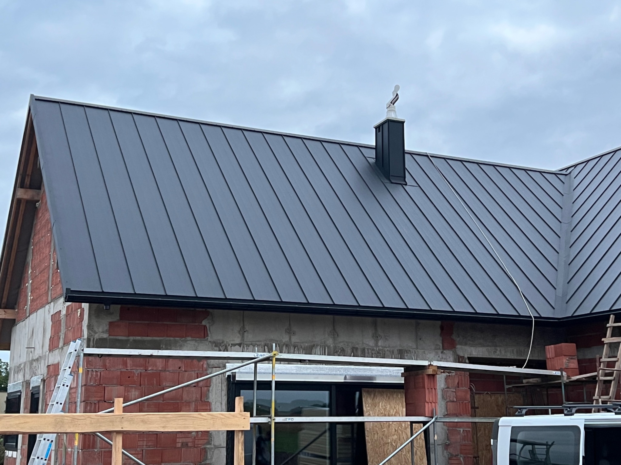 Nowoczesne, grafitowe krycie dachu z blachy na domu w trakcie budowy. Widoczny komin z ozdobną figurką na szczycie i rusztowania.