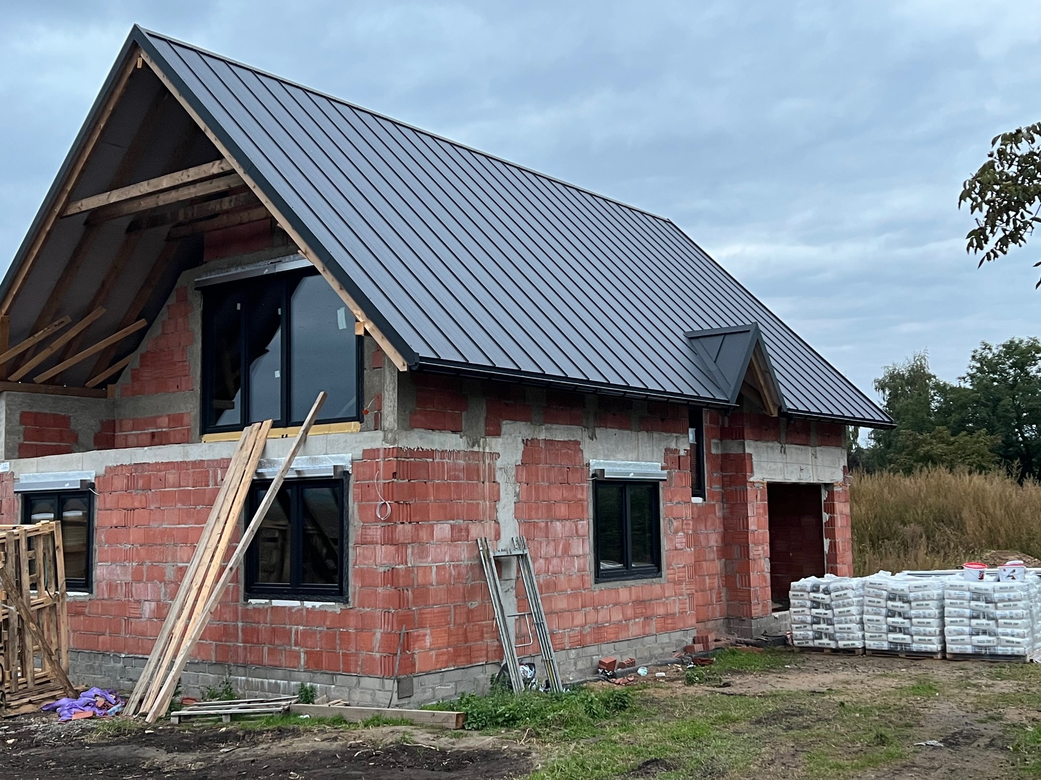 Dom w trakcie budowy z czarnym, metalowym dachem. Widoczne cegły, okna i elementy konstrukcji dachu. Na ziemi leżą materiały budowlane.