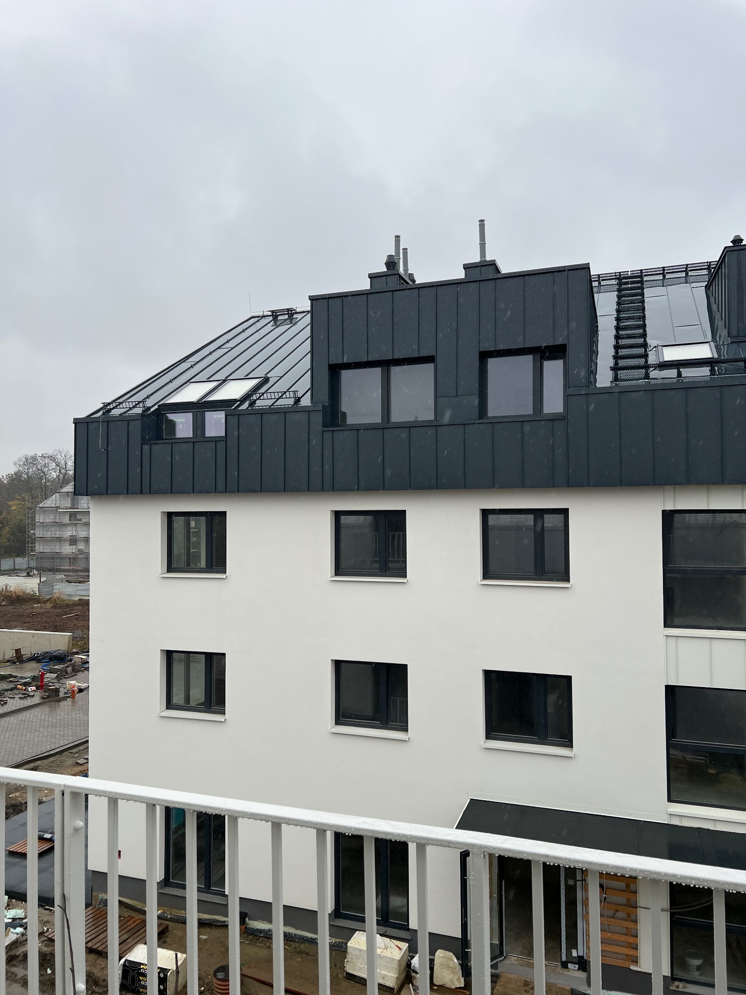 Nowoczesny budynek z dachem z blachy w kolorze grafitowym, widok z balkonu. Na dachu okna dachowe i kominy. W tle plac budowy i pochmurne niebo.