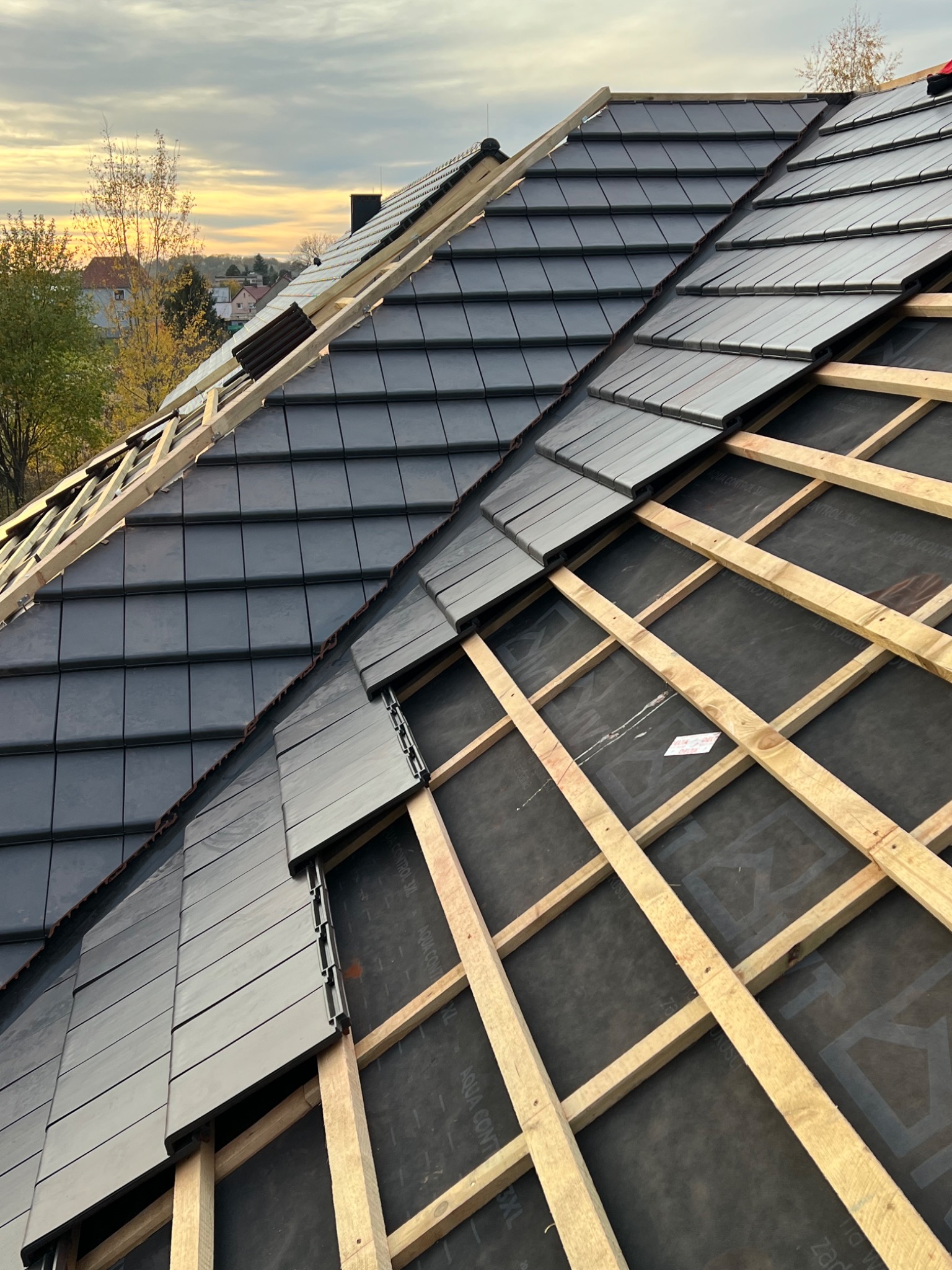 Układanie grafitowej dachówki na drewnianej konstrukcji dachu, widok z góry. Część dachu już pokryta, w tle zabudowa jednorodzinna i zachodzące słońce.