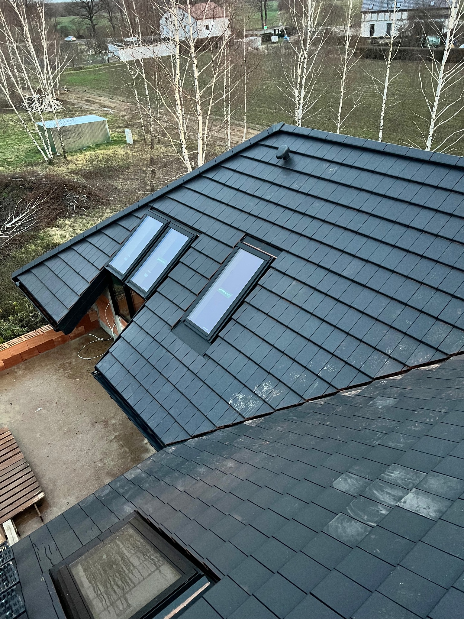 Czarny dach z dachówki ceramicznej z trzema oknami dachowymi, widok z góry na tle drzew i zabudowań. Dach świeżo położony, z widocznymi detalami wykończenia.