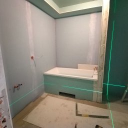 SASIUK SPÓŁKA Z OGRANICZONĄ ODPOWIEDZIALNOŚCIĄ - Łazienka w trakcie prac wykończeniowych; widoczna wanna obudowana płytami gipsowo-kartonowymi, ściany przygotowane pod malowanie, poziomica laserowa wyznacza linie proste, surowy stan wykończenia.