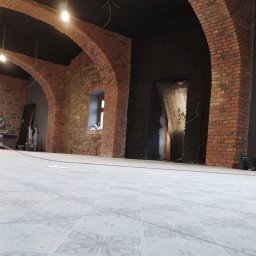 SASIUK SPÓŁKA Z OGRANICZONĄ ODPOWIEDZIALNOŚCIĄ - Wnętrze w trakcie prac wykończeniowych z widocznymi ceglanymi łukami, surowymi kamiennymi ścianami, nową podłogą w jasne wzory i prowizorycznym oświetleniem