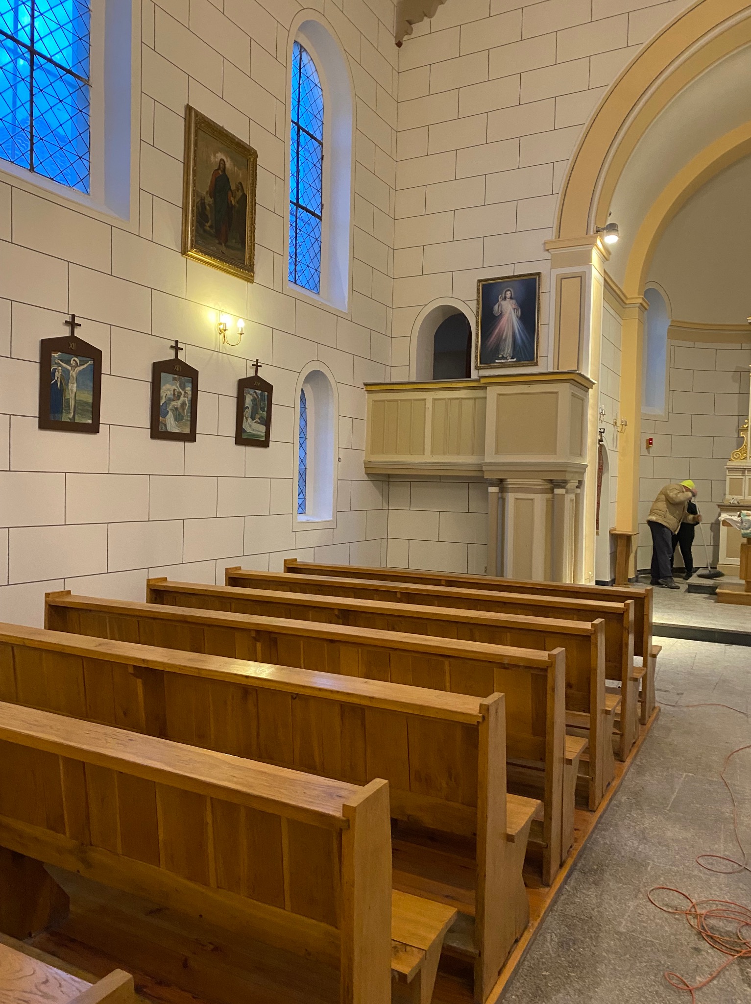 Wnętrze kościoła z drewnianymi ławkami, białymi ścianami z cegieł, obrazami religijnymi i osobą sprzątającą w tle.