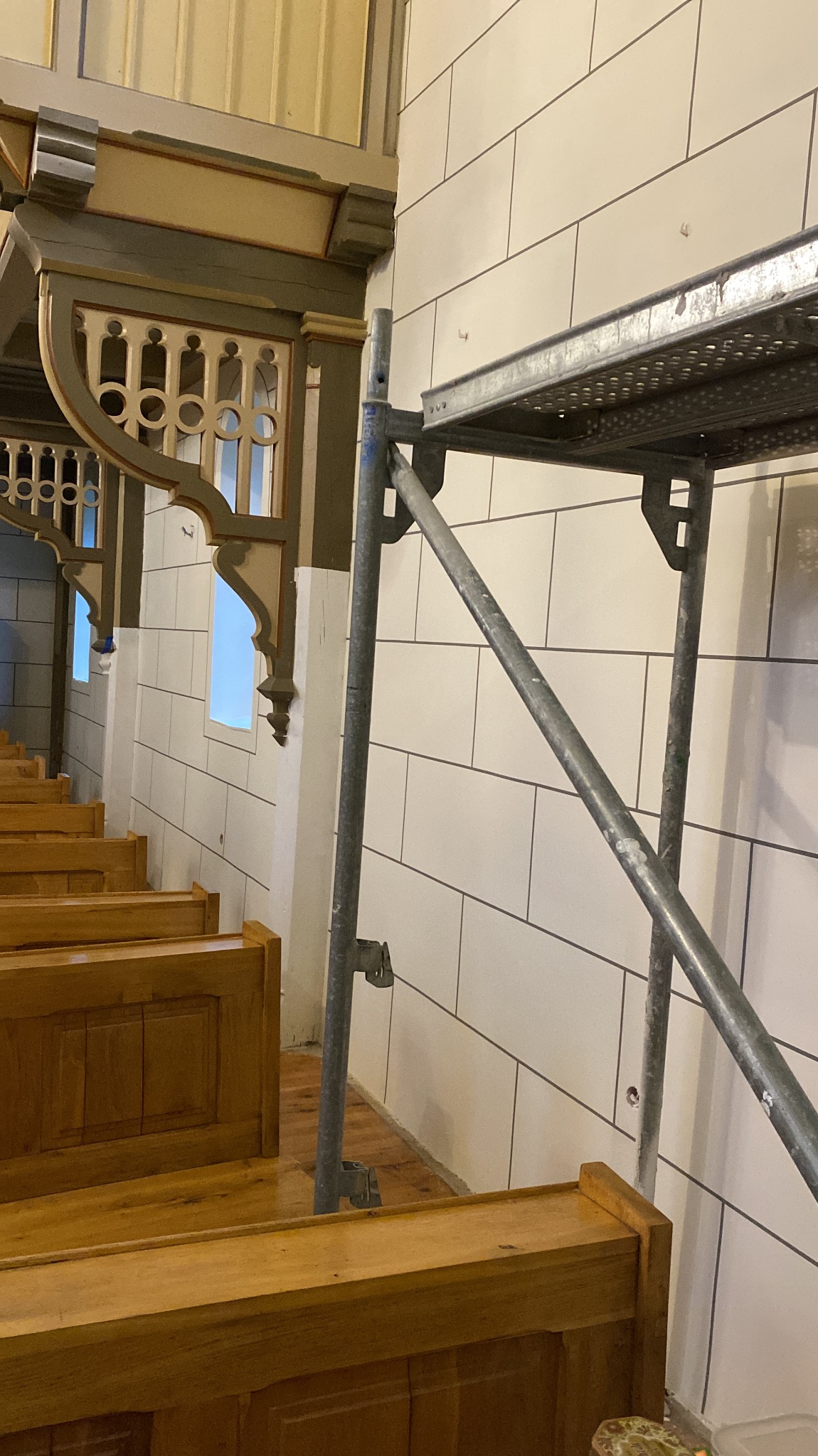 Wnętrze kościoła w trakcie prac: białe, prostokątne płytki na ścianie, drewniane ławki, ozdobne, drewniane elementy architektoniczne i metalowe rusztowanie.