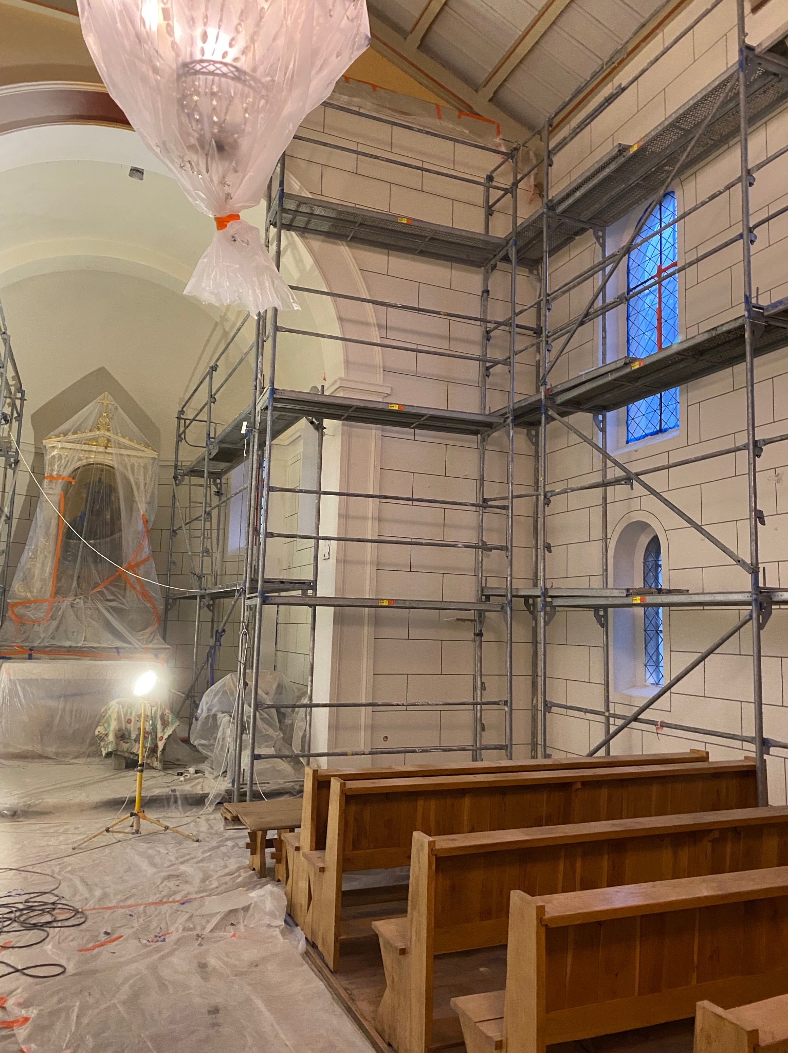 Wnętrze kościoła w trakcie prac: rusztowania, zabezpieczona folią lampa i ołtarz, ławki, podłoga przykryta folią ochronną, widoczne ślady prac.