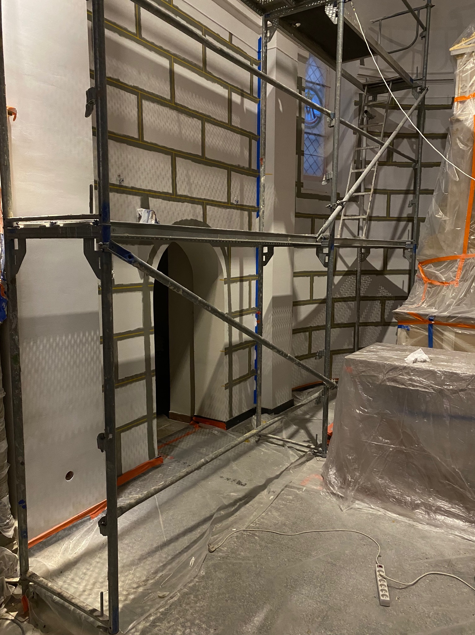 Wnętrze w trakcie remontu, ściany z nałożonymi płytami kartonowo-gipsowymi imitującymi cegłę, widoczne rusztowania i zabezpieczenia folią.