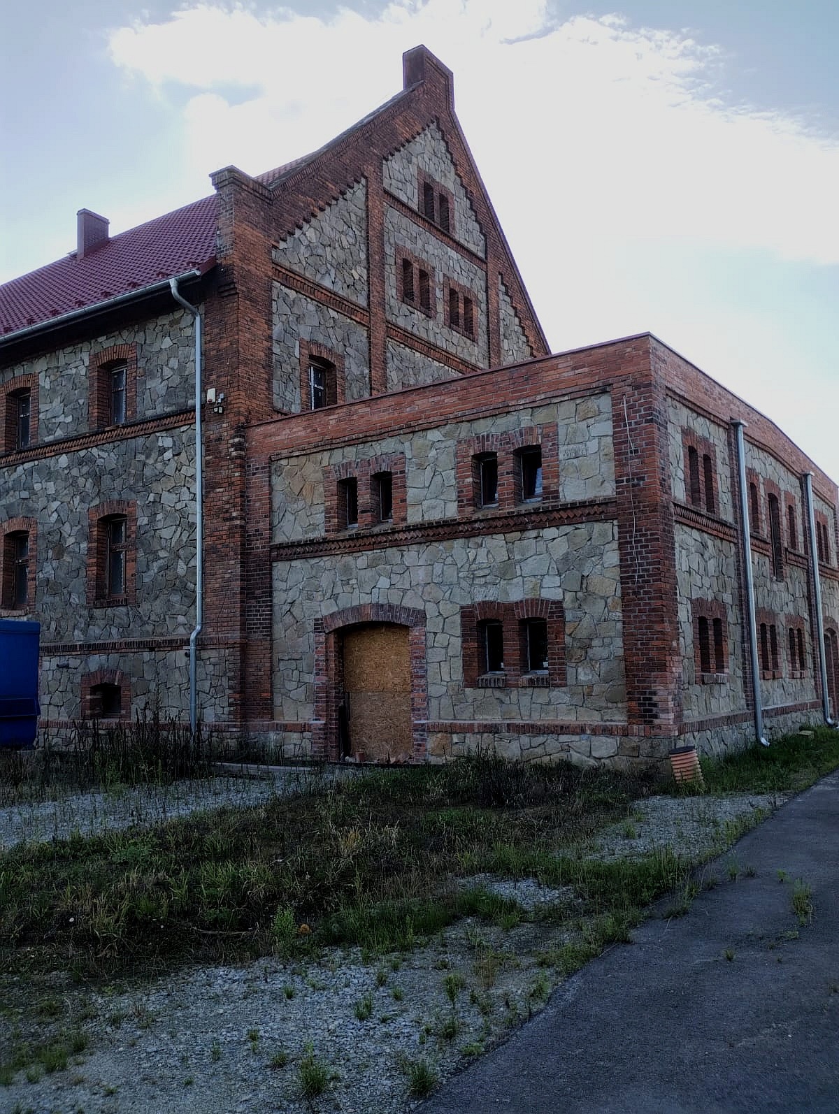 Dwukondygnacyjny budynek z kamienia i czerwonej cegły, z nowym czerwonym dachem, okna zabite deskami, otoczony zaniedbaną zielenią.