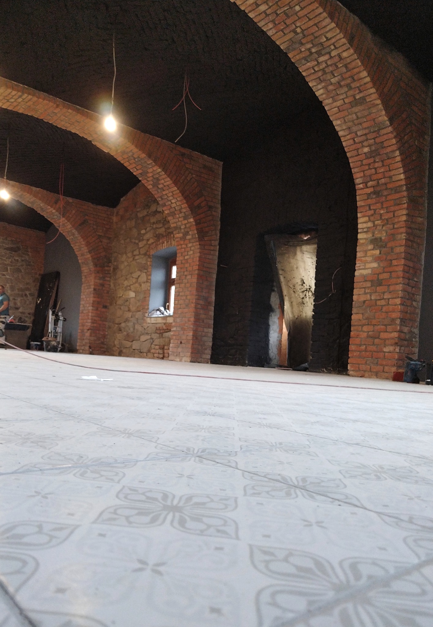 Wnętrze w trakcie prac wykończeniowych z widocznymi ceglanymi łukami, surowymi kamiennymi ścianami, nową podłogą w jasne wzory i prowizorycznym oświetleniem