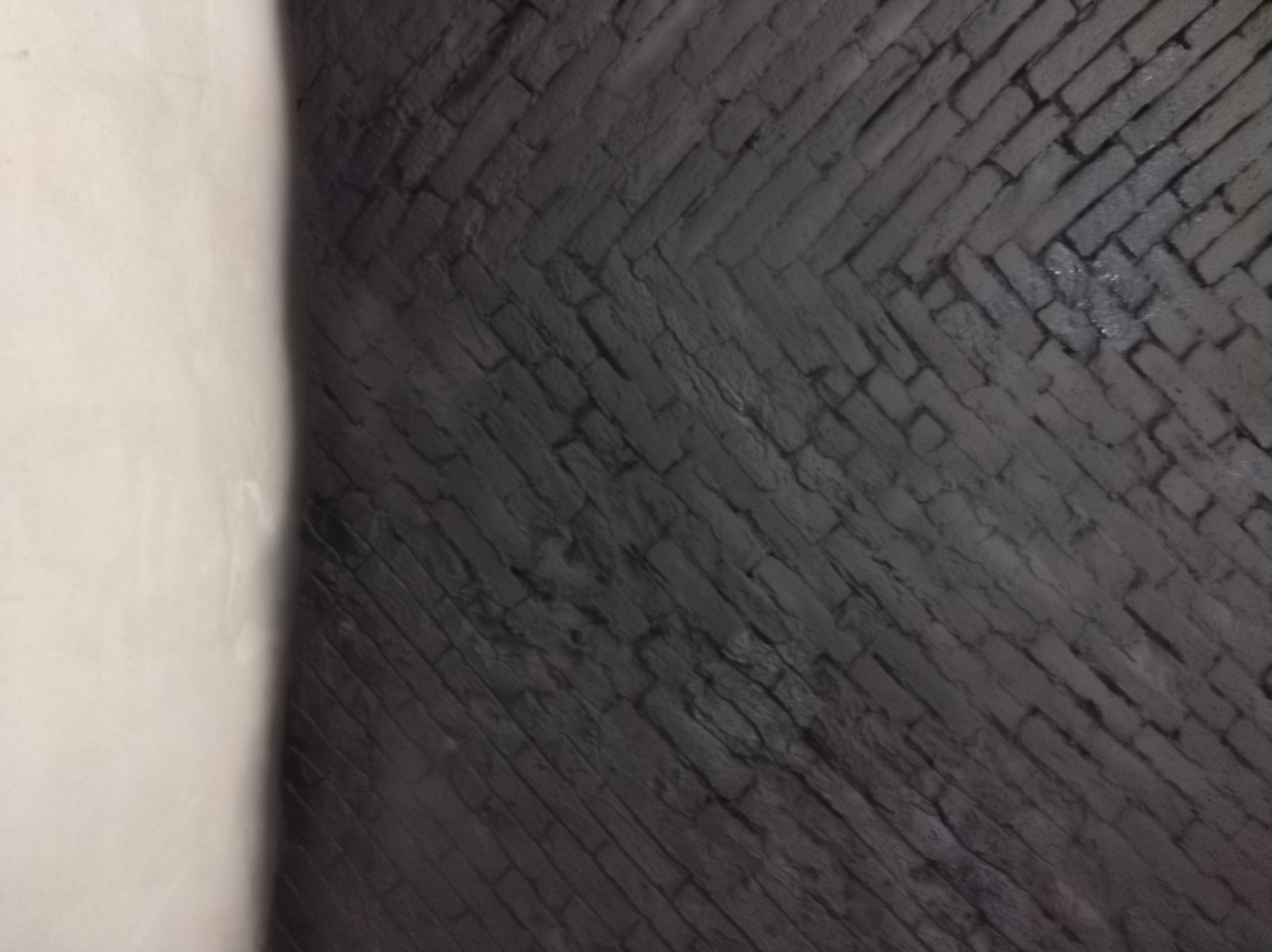 Ciemnoszara ściana z cegieł ułożonych w jodełkę, częściowo przysłonięta jasną ścianą.