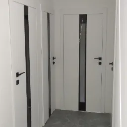 Korytarz z trzema parami białych drzwi z czarnymi wstawkami i czarnymi klamkami, szare marmurowe płytki na podłodze, minimalistyczne oświetlenie sufitowe.