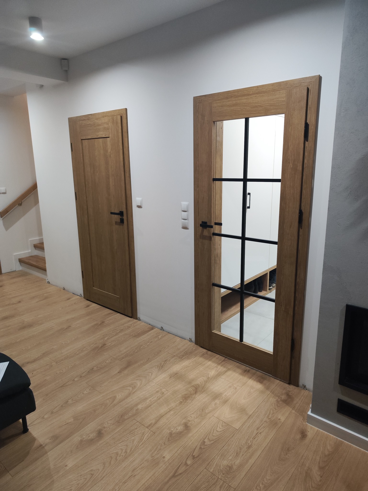 Dwa rodzaje drzwi wewnętrznych w nowoczesnym mieszkaniu: pełne drzwi drewniane z czarną klamką oraz drzwi szklane z czarnymi szprosami, ukazujące wnętrze szafy z butami.