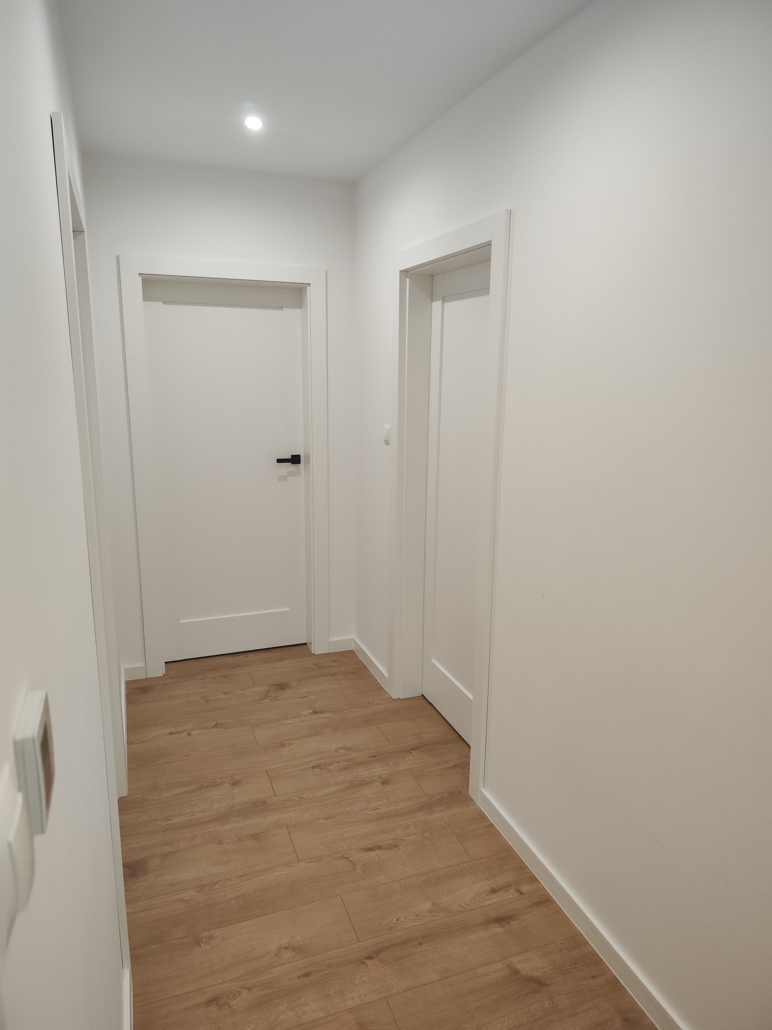 Korytarz z dwiema białymi, zamkniętymi drzwiami z czarnymi klamkami i panelami podłogowymi w odcieniach brązu.