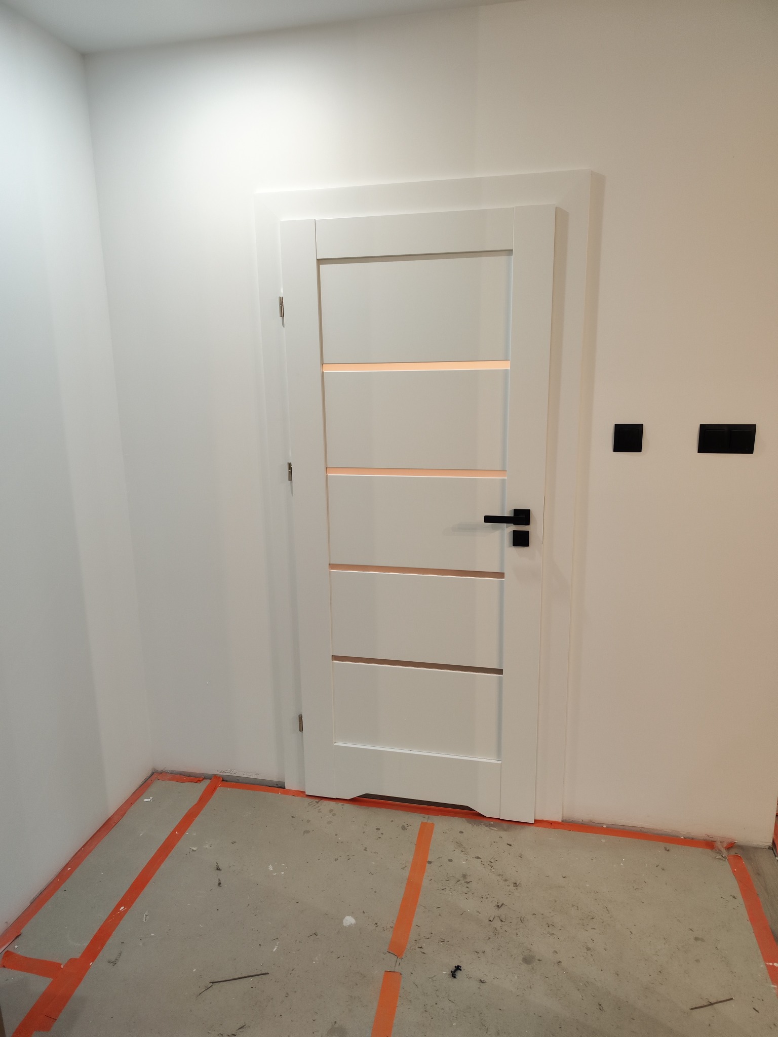 Białe drzwi wewnętrzne z czterema poziomymi przeszkleniami, czarna klamka, widoczne przygotowanie podłoża z taśmą malarską na podłodze, białe ściany, czarne włączniki światła.