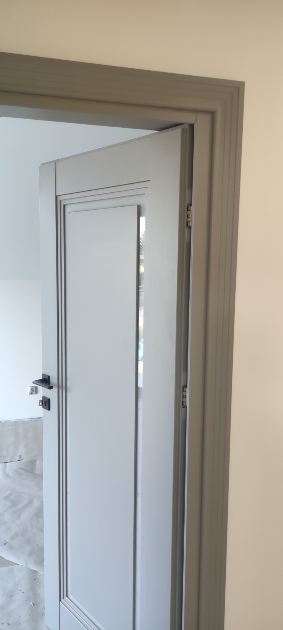 Otwarta, szara, wewnętrzna płycinowa drzwi z czarną klamką i widocznymi zawiasami, osadzona w szarej futrynie, z widoczną podłogą zabezpieczoną folią malarską.