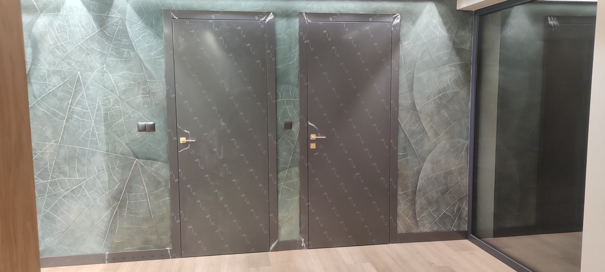 Dwa szare, metalowe skrzydła drzwiowe z złotymi klamkami, zabezpieczone folią ochronną, na tle ściany z motywem liści i szklanej ściany.