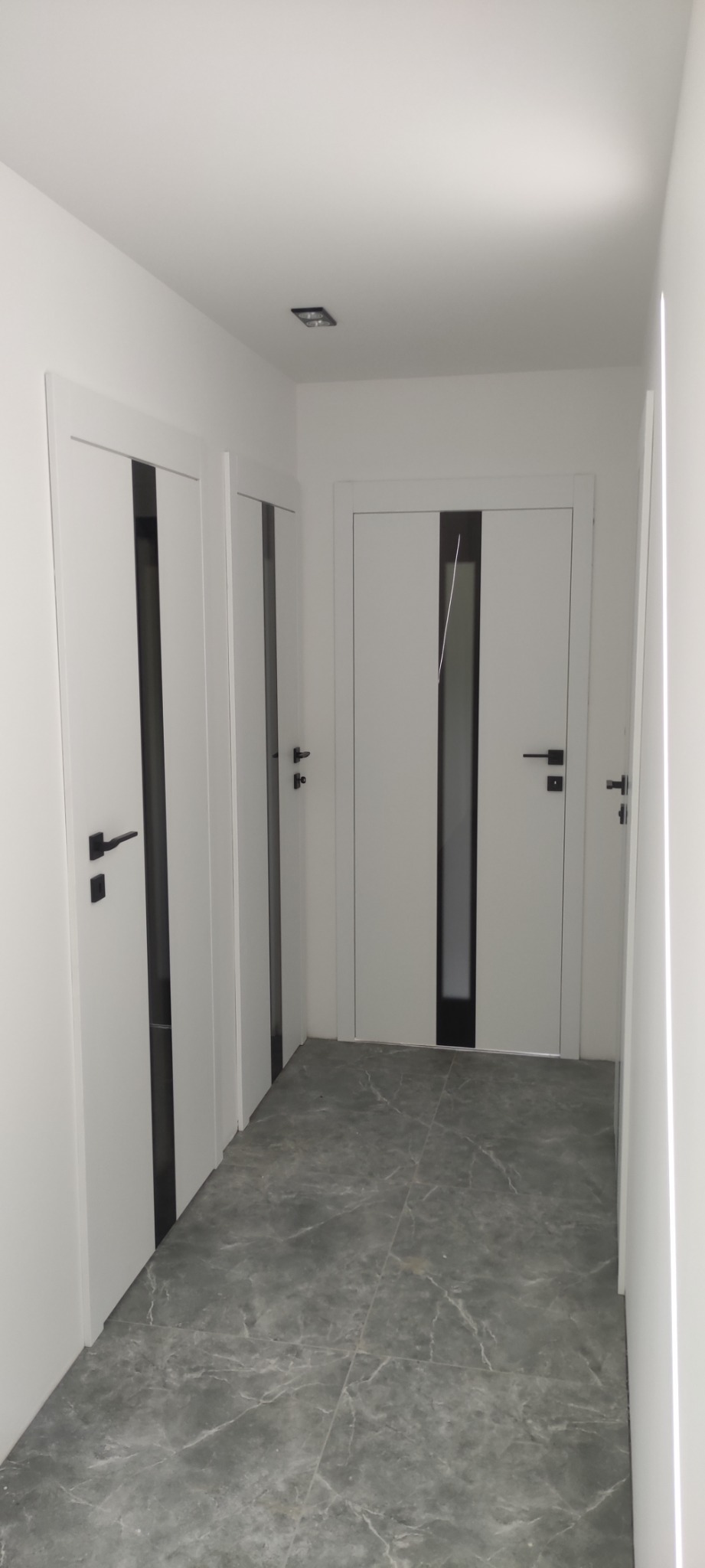 Korytarz z trzema parami białych drzwi z czarnymi wstawkami i czarnymi klamkami, szare marmurowe płytki na podłodze, minimalistyczne oświetlenie sufitowe.