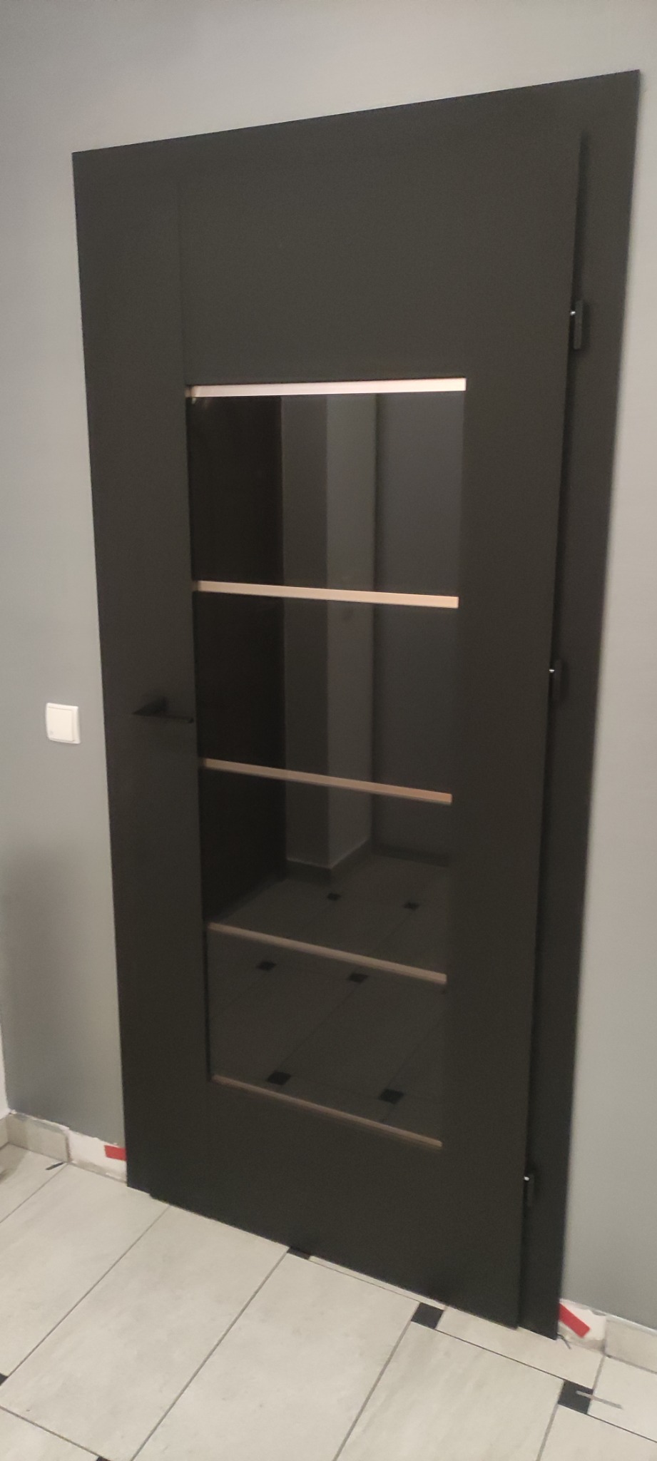 Ciemne drzwi z przeszkleniami i poziomymi szprosami, widoczne na tle szarej ściany i jasnej podłogi z ciemnymi akcentami.