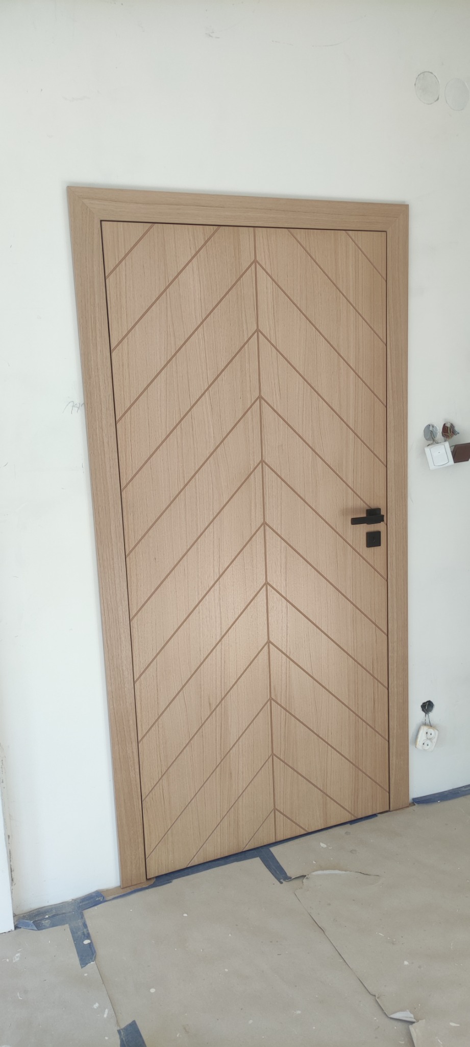 Drewniane drzwi wewnętrzne z geometrycznym wzorem w jodełkę, czarna klamka, widoczne niedoskonałości wykończenia ściany i podłogi.