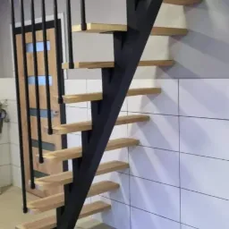 Nowoczesne schody metalowe z drewnianymi stopniami, czarna konstrukcja, minimalistyczny design, widok z dołu, białe płytki na ścianie w tle.