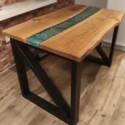 Stół z blatem z naturalnego drewna z żywicą epoksydową w kolorze turkusowym, osadzony na czarnych, metalowych nogach o geometrycznym kształcie, na tle ceglanej ściany i drewnianej podłogi.