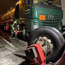 Pracownik w odblaskowym ubraniu wymienia koło w zielonej ciężarówce cysternie WĘGLOSTAL przy użyciu elektrycznego klucza udarowego, obok leży zdemontowane koło i czerwona wyważarka do kół, nocna...