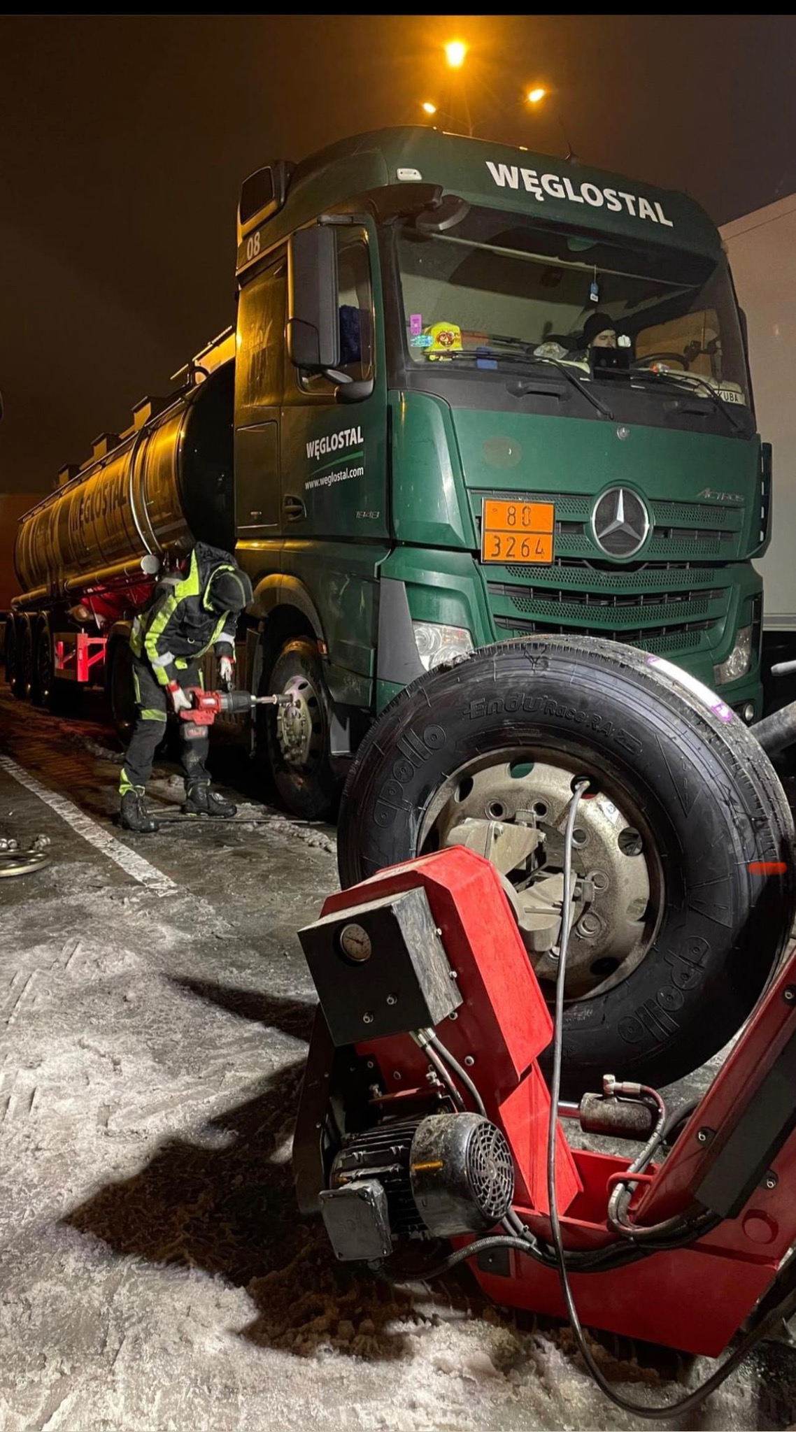 Pracownik w odblaskowym ubraniu wymienia koło w zielonej ciężarówce cysternie WĘGLOSTAL przy użyciu elektrycznego klucza udarowego, obok leży zdemontowane koło i czerwona wyważarka do kół, nocna...