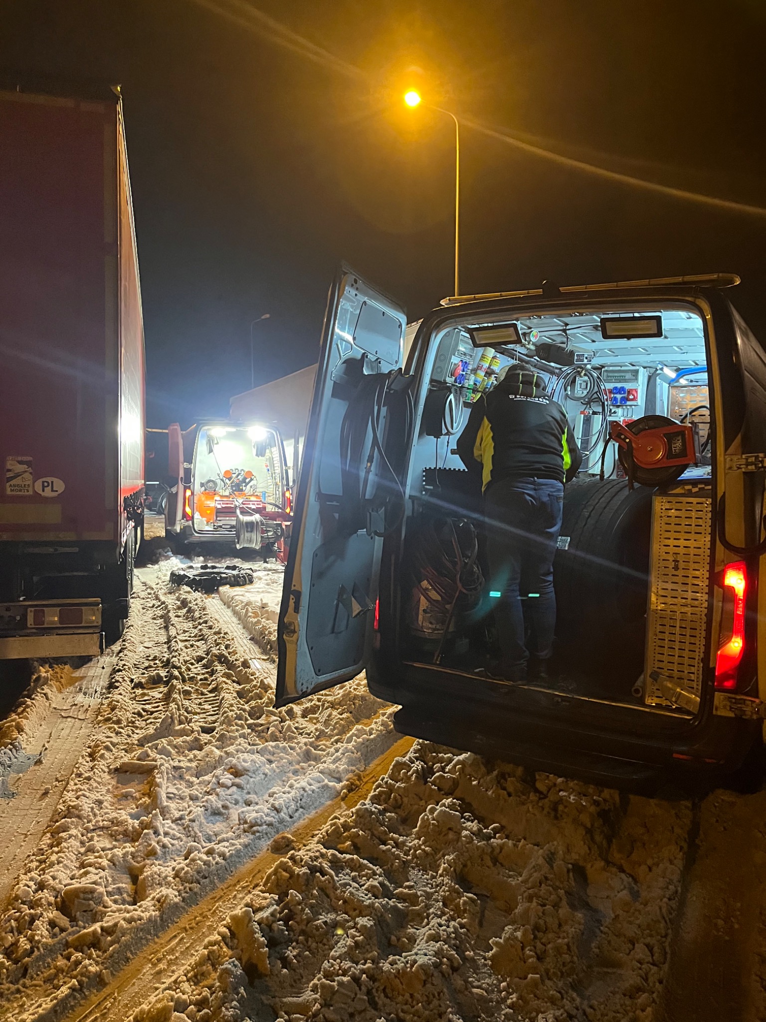 Mechanik w pojeździe serwisowym przy drodze, prawdopodobnie wymiana opon ciężarówki w warunkach zimowych, widoczne narzędzia i opony wewnątrz pojazdu, śnieg na poboczu.