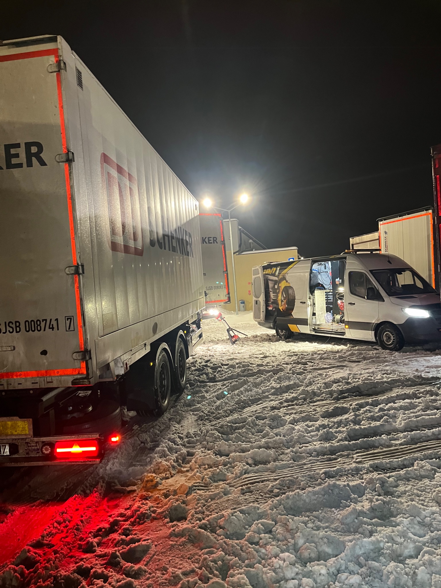 Ciężarówka z naczepą marki Schenker stoi na parkingu pokrytym śniegiem obok białego busa z otwartymi drzwiami, w którym widać narzędzia i wyposażenie warsztatowe. Na boku busa widnieje grafika opon...