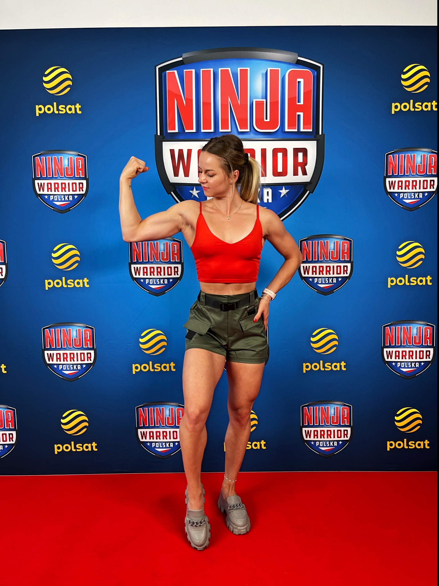 Uśmiechnięta wysportowana kobieta w czerwonym topie i zielonych szortach prezentuje bicepsy na tle ściany z logotypami Ninja Warrior Polska i Polsat.