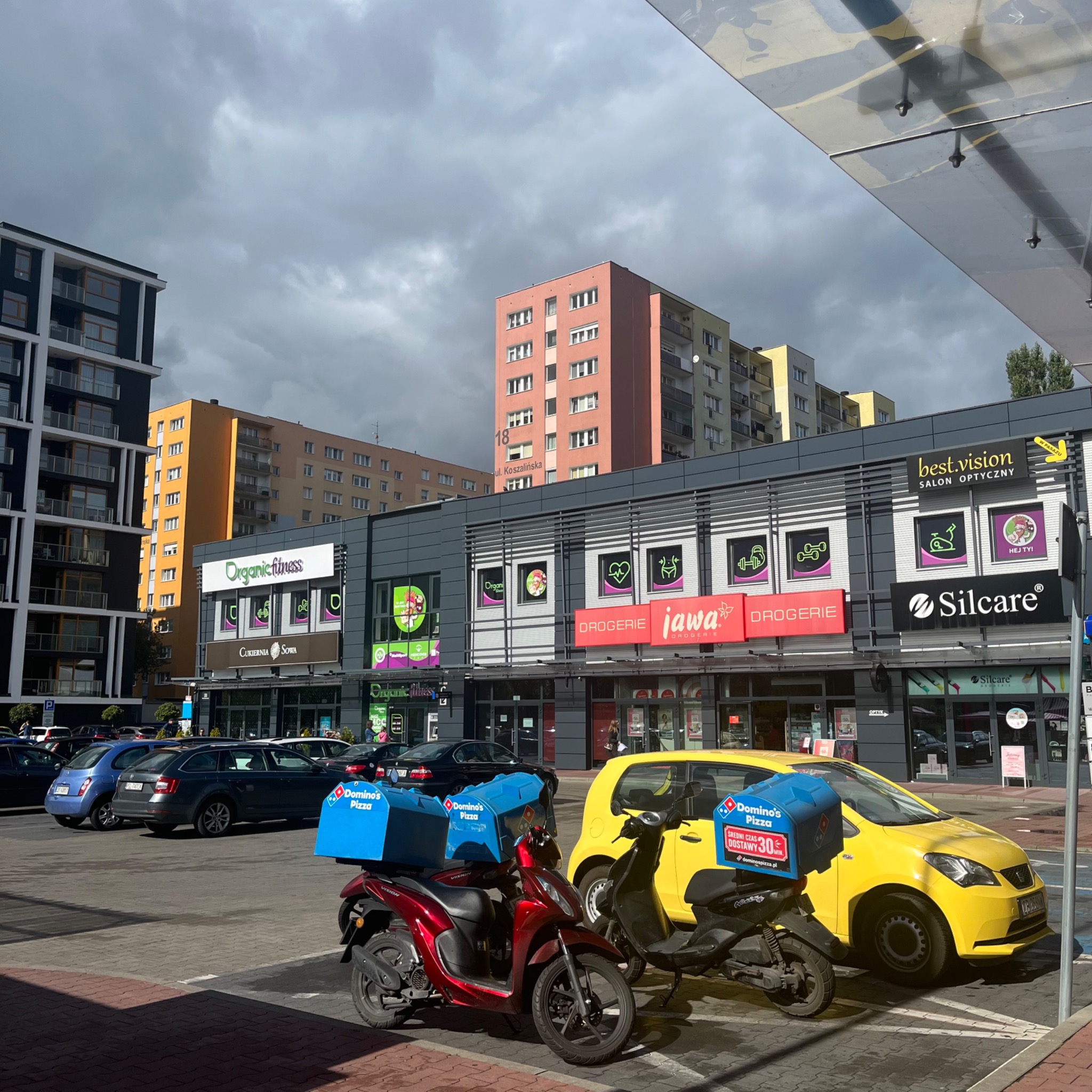 Dwa skutery dostawcze Domino's Pizza zaparkowane przed budynkiem z lokalami usługowymi, w tym Organic Fitness i salonem optycznym Best Vision.