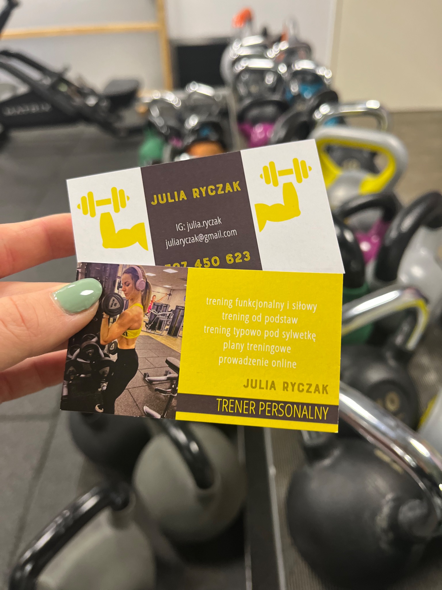 Wizytówka trenera personalnego Julia Ryczak na tle sprzętu do ćwiczeń, prezentująca dane kontaktowe i zakres usług: trening funkcjonalny, siłowy, dopasowany do sylwetki, plany treningowe...
