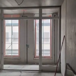 Surowy stan wykończenia wnętrza z zamontowanymi oknami i przygotowanym otworem na drzwi; widoczne kable, narzędzia i surowe ściany betonowe.