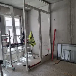 Montaż szklanej konstrukcji w surowym wnętrzu, widoczne rusztowanie, poziomica i odblaskowa kamizelka, surowy beton na ścianach i podłodze.