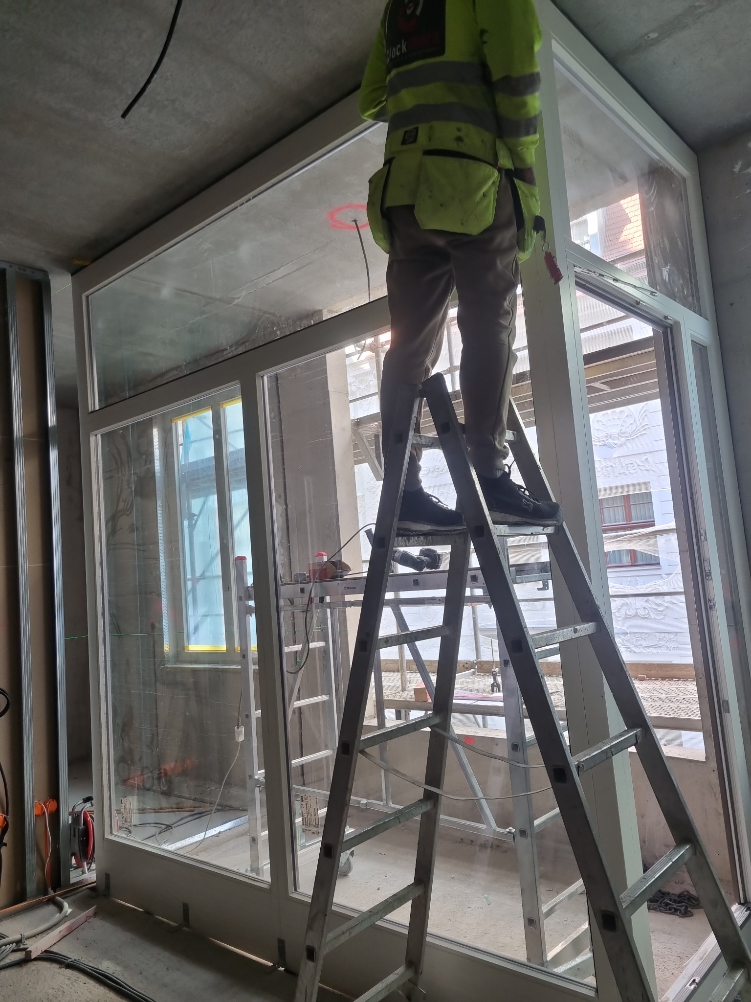 Pracownik w odblaskowej kamizelce, stojący na drabinie, montuje duże okno z białymi ramami w surowym wnętrzu budynku w trakcie budowy. Widoczne narzędzia i materiały budowlane.