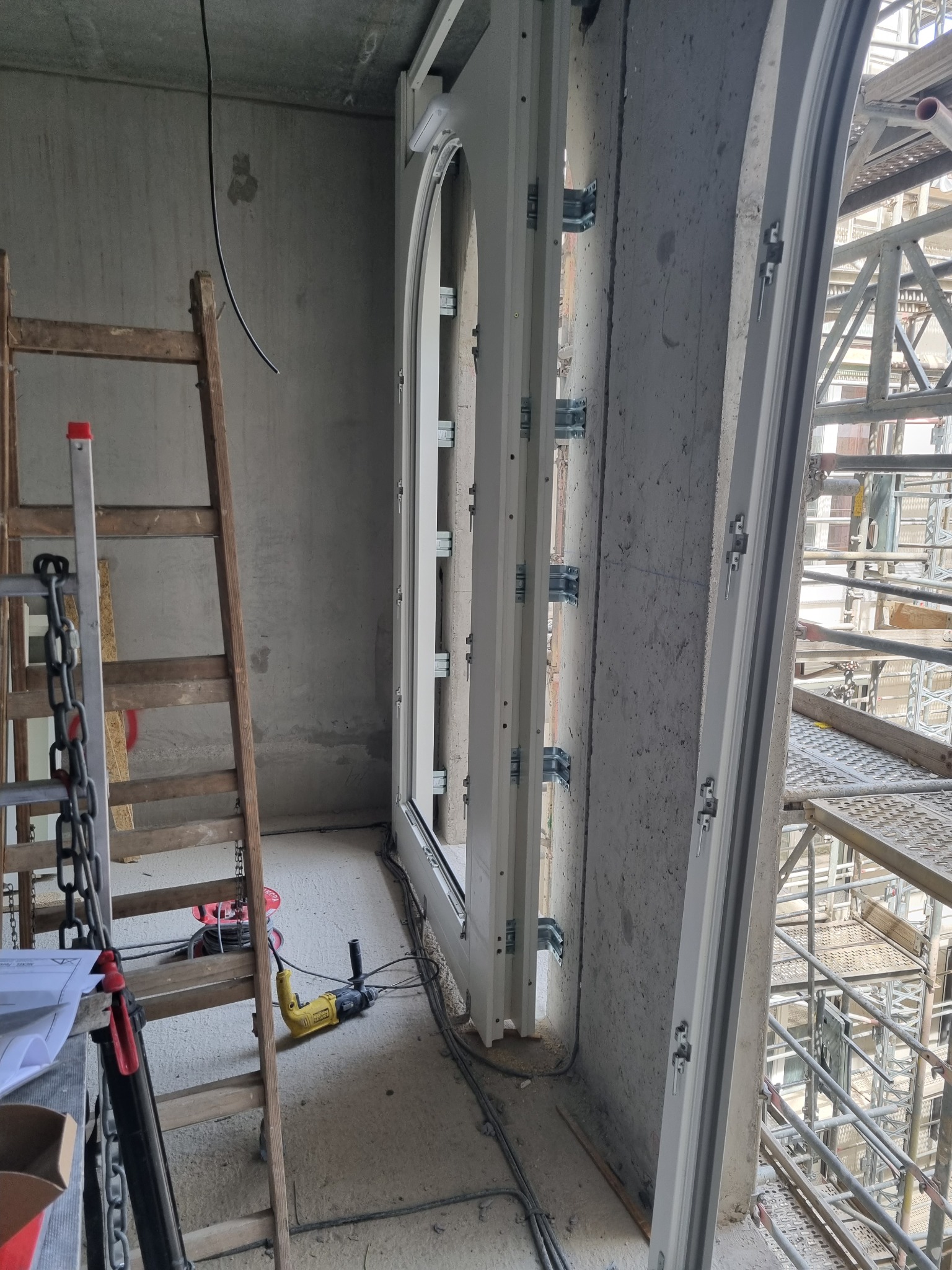 Montaż łukowych drzwi w surowym stanie, widoczne narzędzia i rusztowanie za oknem.