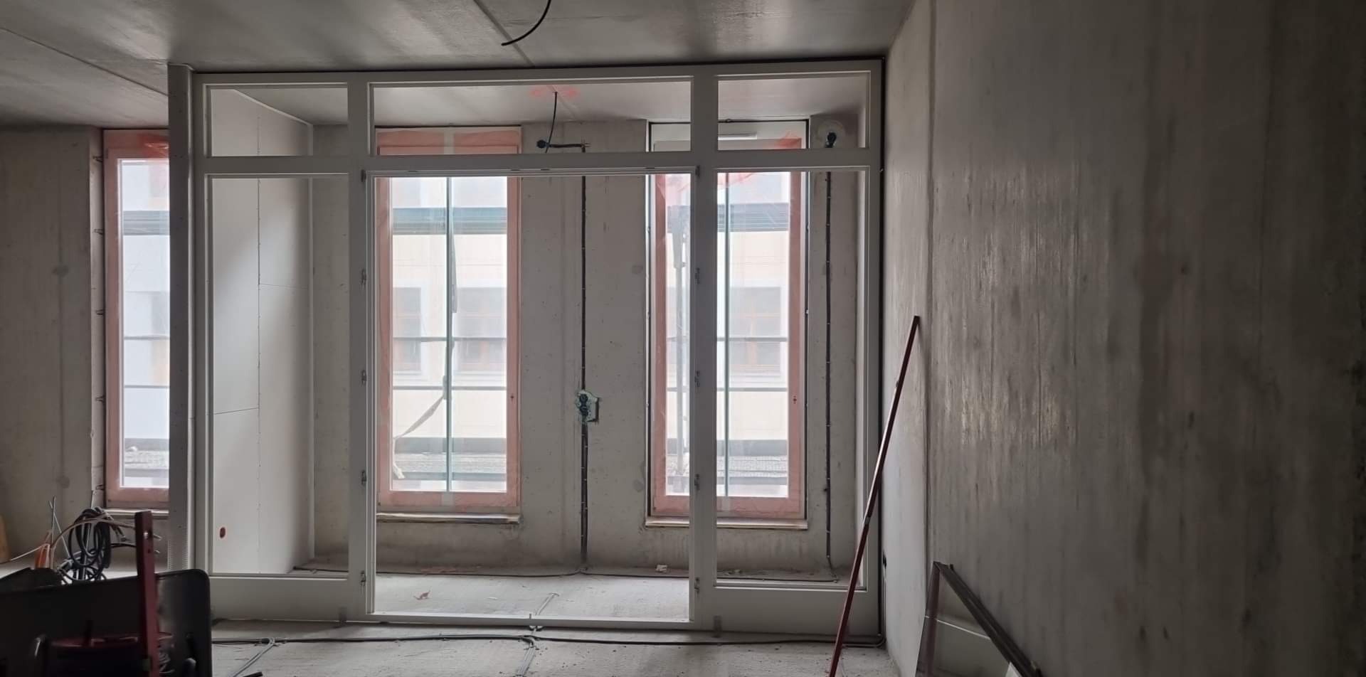 Surowy stan wykończenia wnętrza z zamontowanymi oknami i przygotowanym otworem na drzwi; widoczne kable, narzędzia i surowe ściany betonowe.