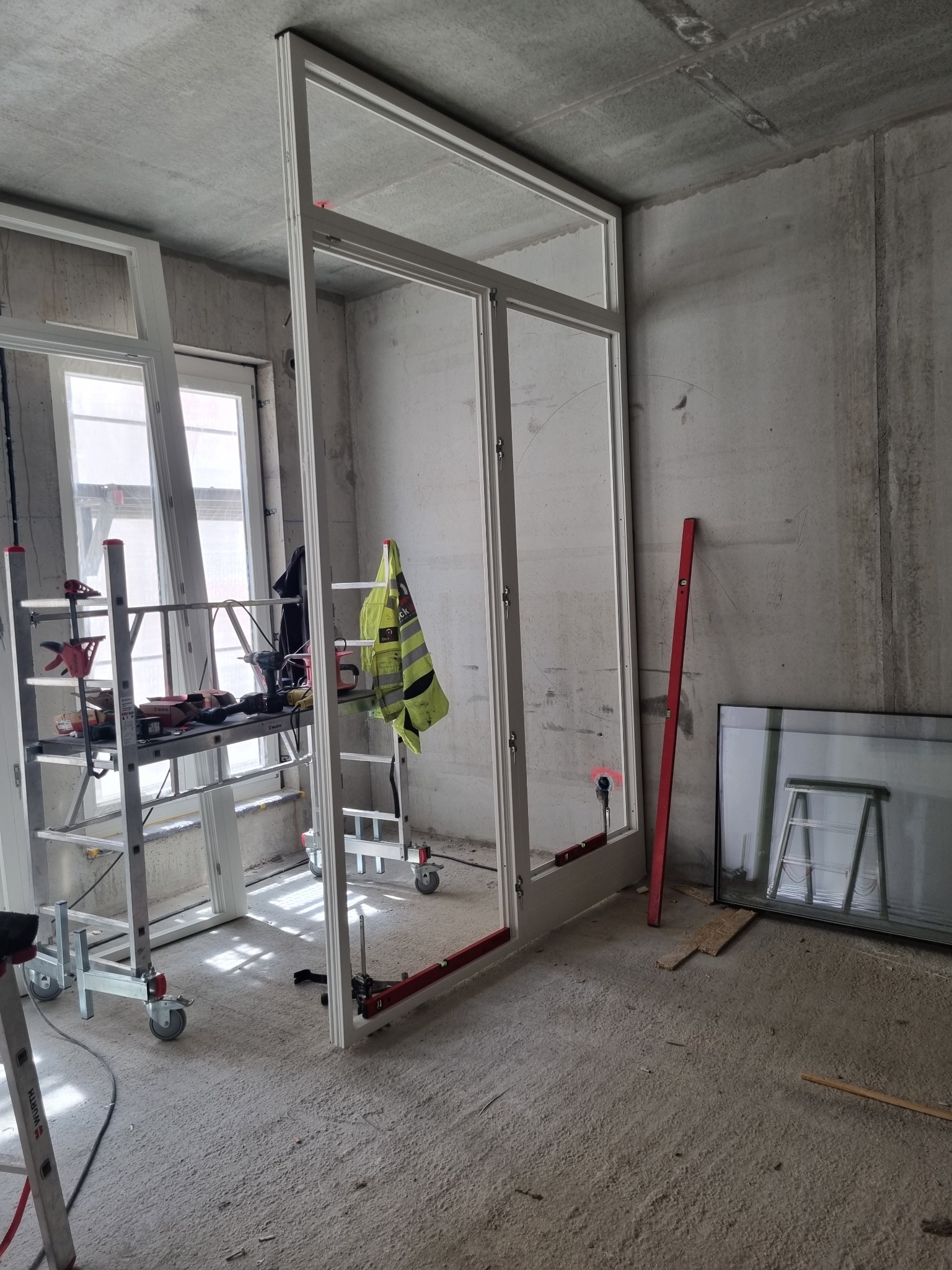 Montaż szklanej konstrukcji w surowym wnętrzu, widoczne rusztowanie, poziomica i odblaskowa kamizelka, surowy beton na ścianach i podłodze.