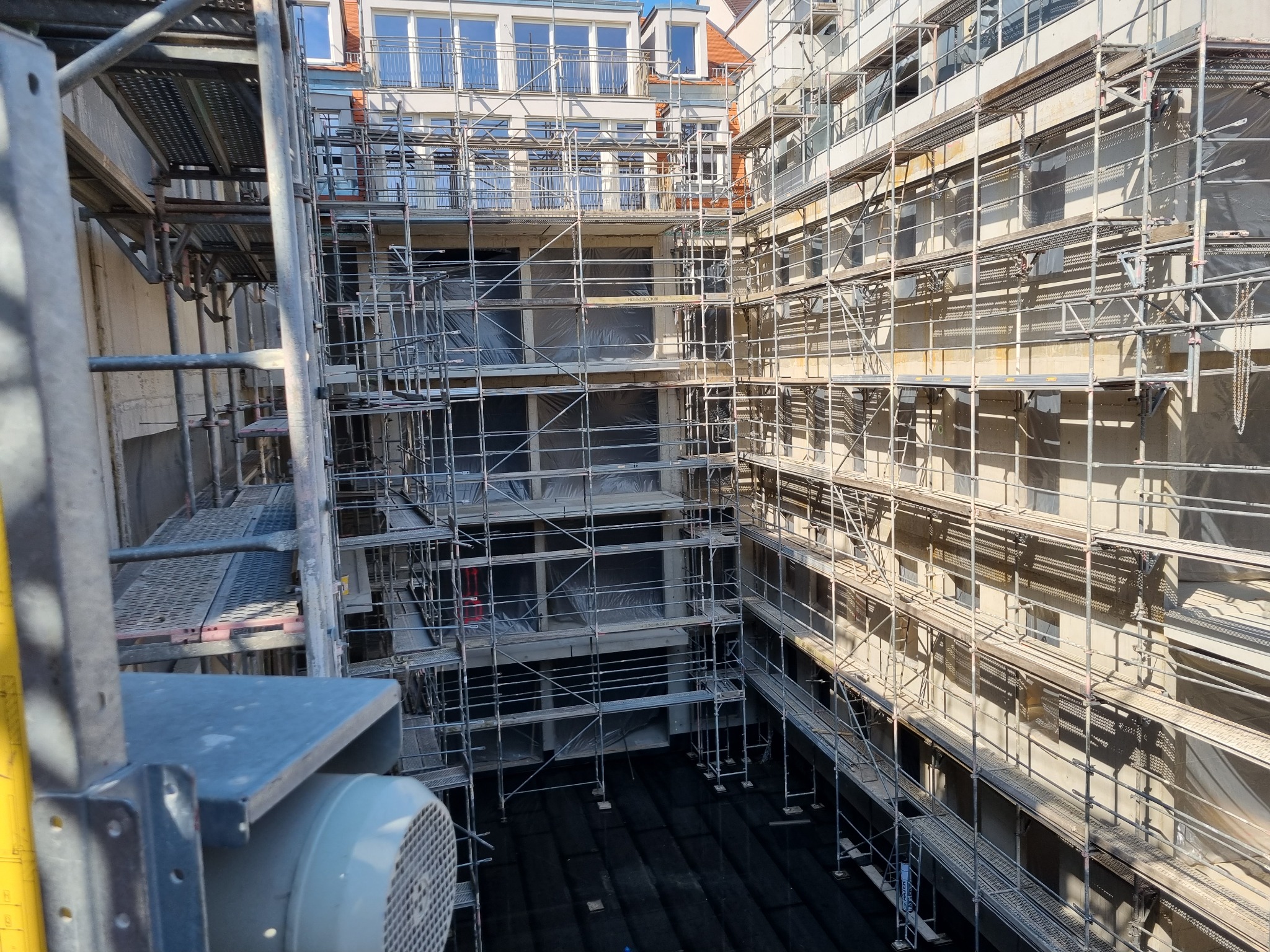 Budynek w trakcie budowy, otoczony rusztowaniami, z widocznymi oknami zabezpieczonymi folią, ujęcie z góry z fragmentem maszyny budowlanej w kadrze.