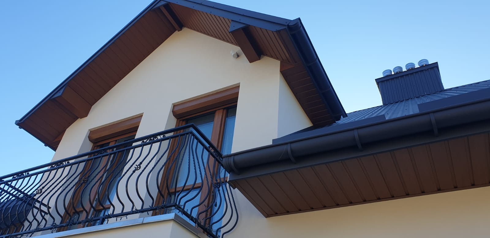 Dwukondygnacyjny dom z beżową elewacją, ciemnobrązową podbitką, dachem z blachy i balkonem z ozdobną, metalową balustradą.